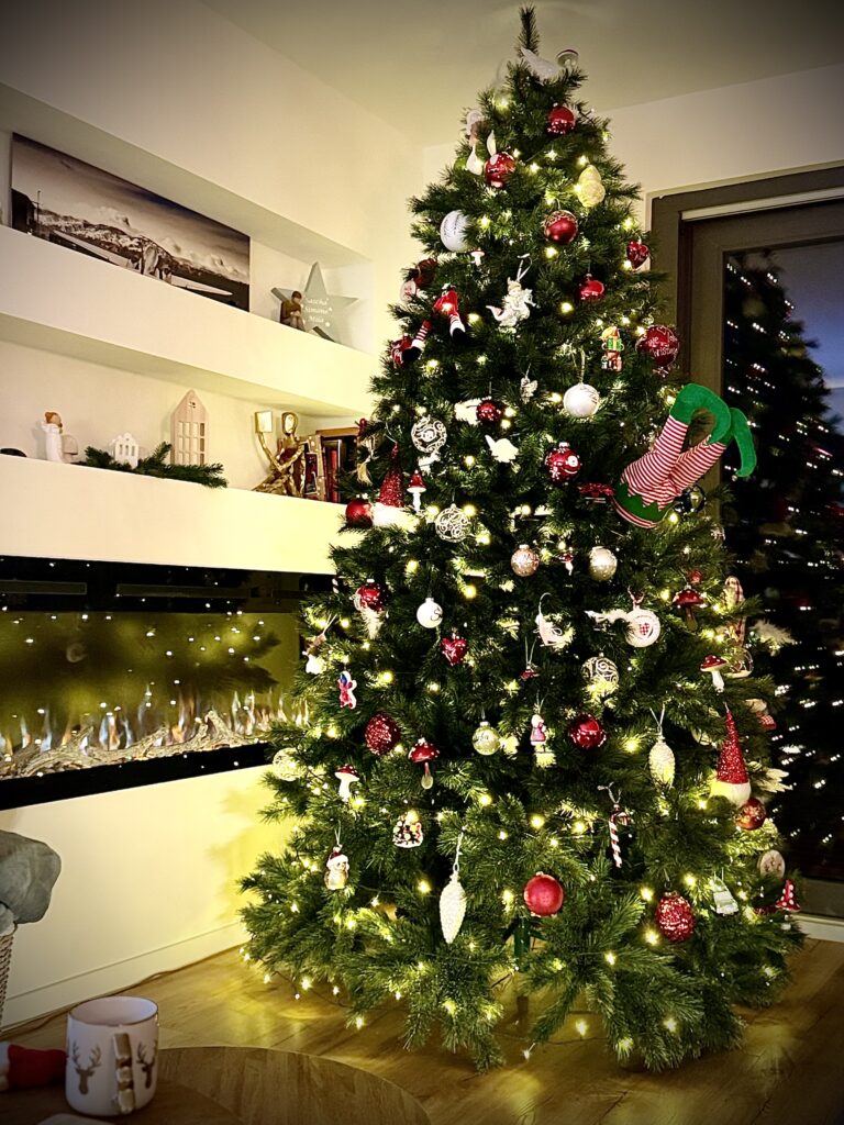 Geschmückter Weihnachtsbaum mit Lichterkette und roten Kugeln neben einem Kamin