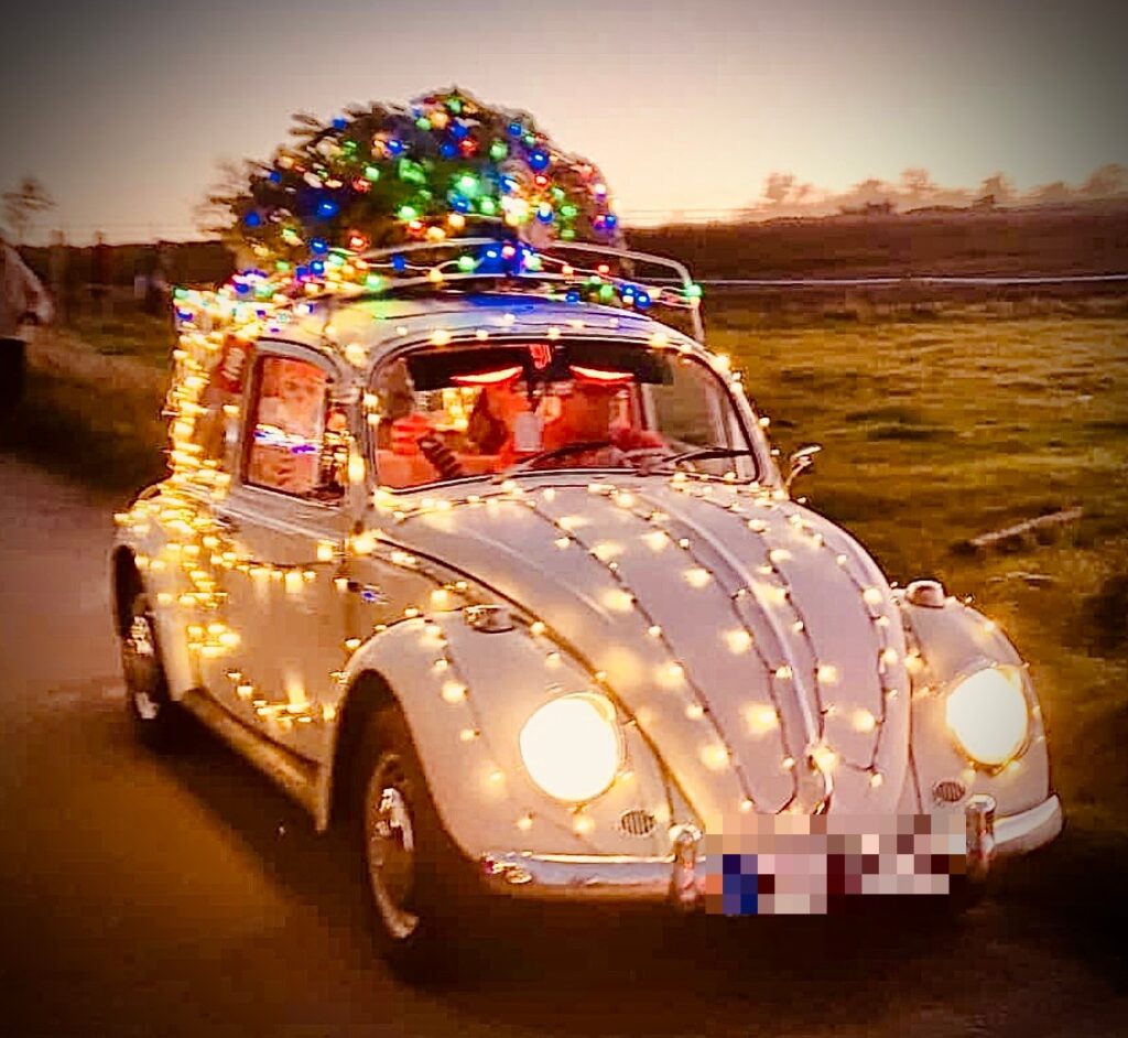Weißer VW Käfer mit Lichterketten und einem leuchtenden Weihnachtsbaum auf dem Dach